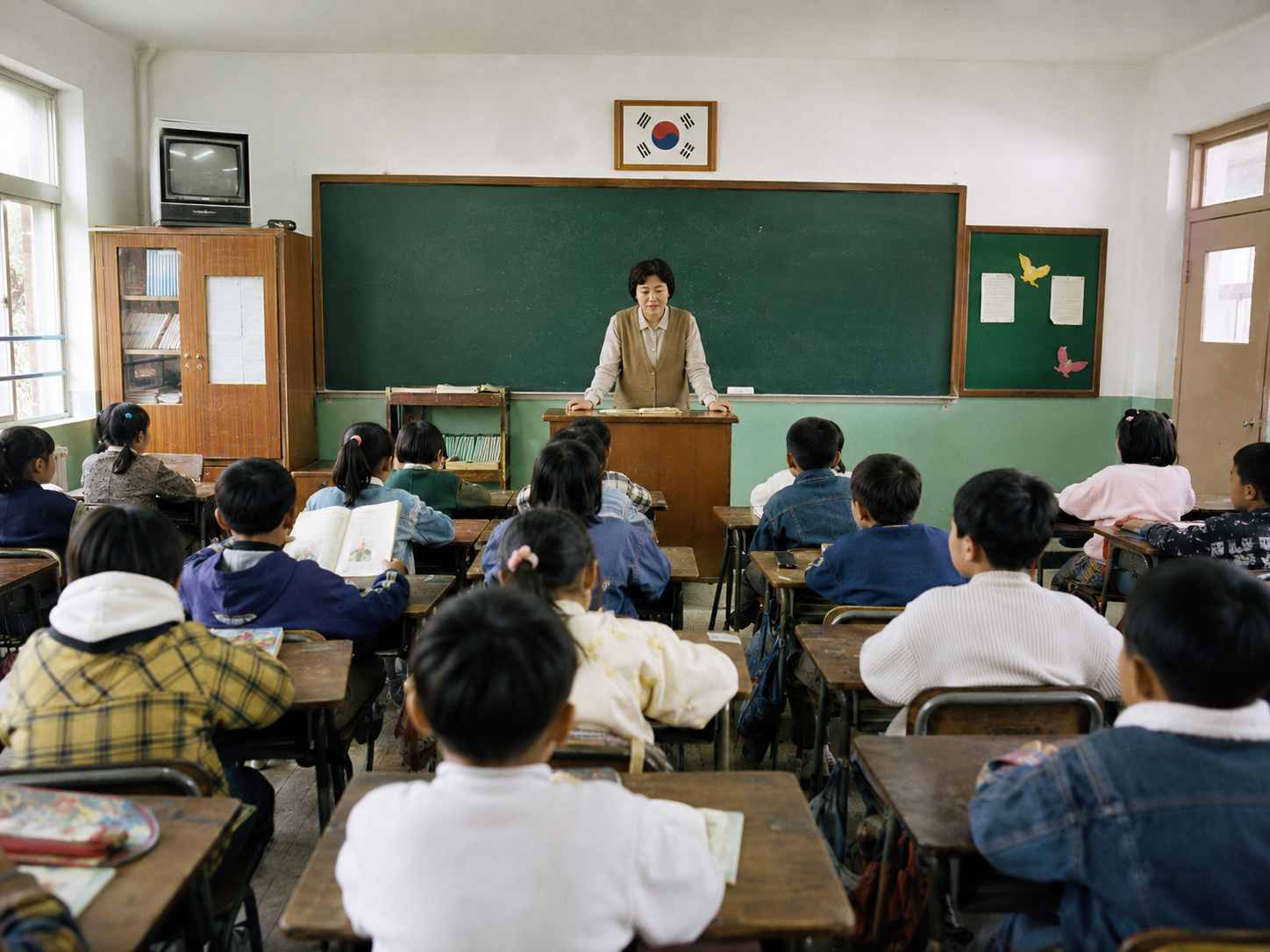 녹색 칠판과 나무 책상이 놓인 오래된 교실에서 학생들이 앞을 바라보며 수업을 듣는 모습