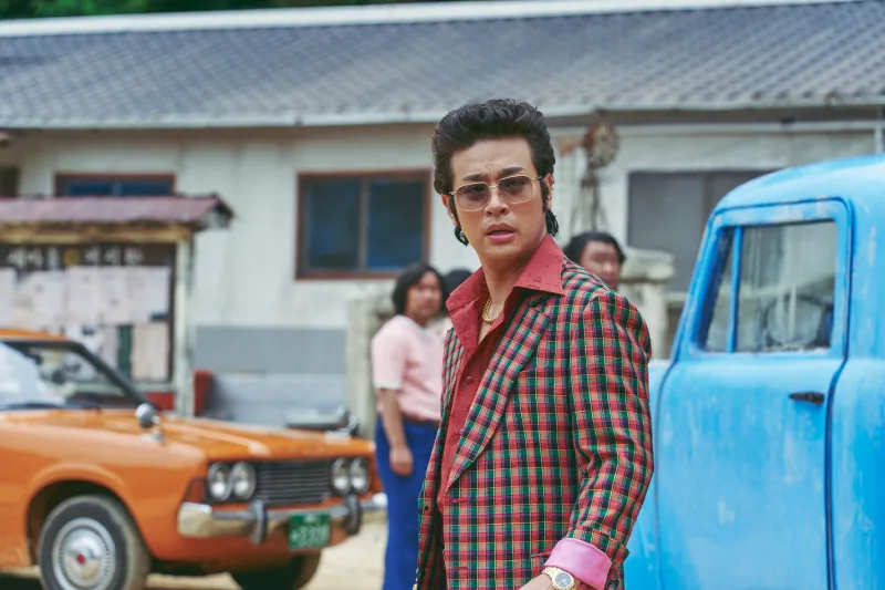 어느 어촌을 배경으로 빨강 슈트에 검은 썬글라스를 끼고 머리를 넘긴 영화 밀수의 박정민