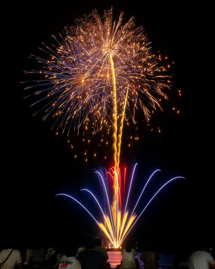 밤하늘을 수놓은 후쿠오카의 화려한 불꽃놀이, 바닷가에 앉은 사람들의 뒷모습과 함께