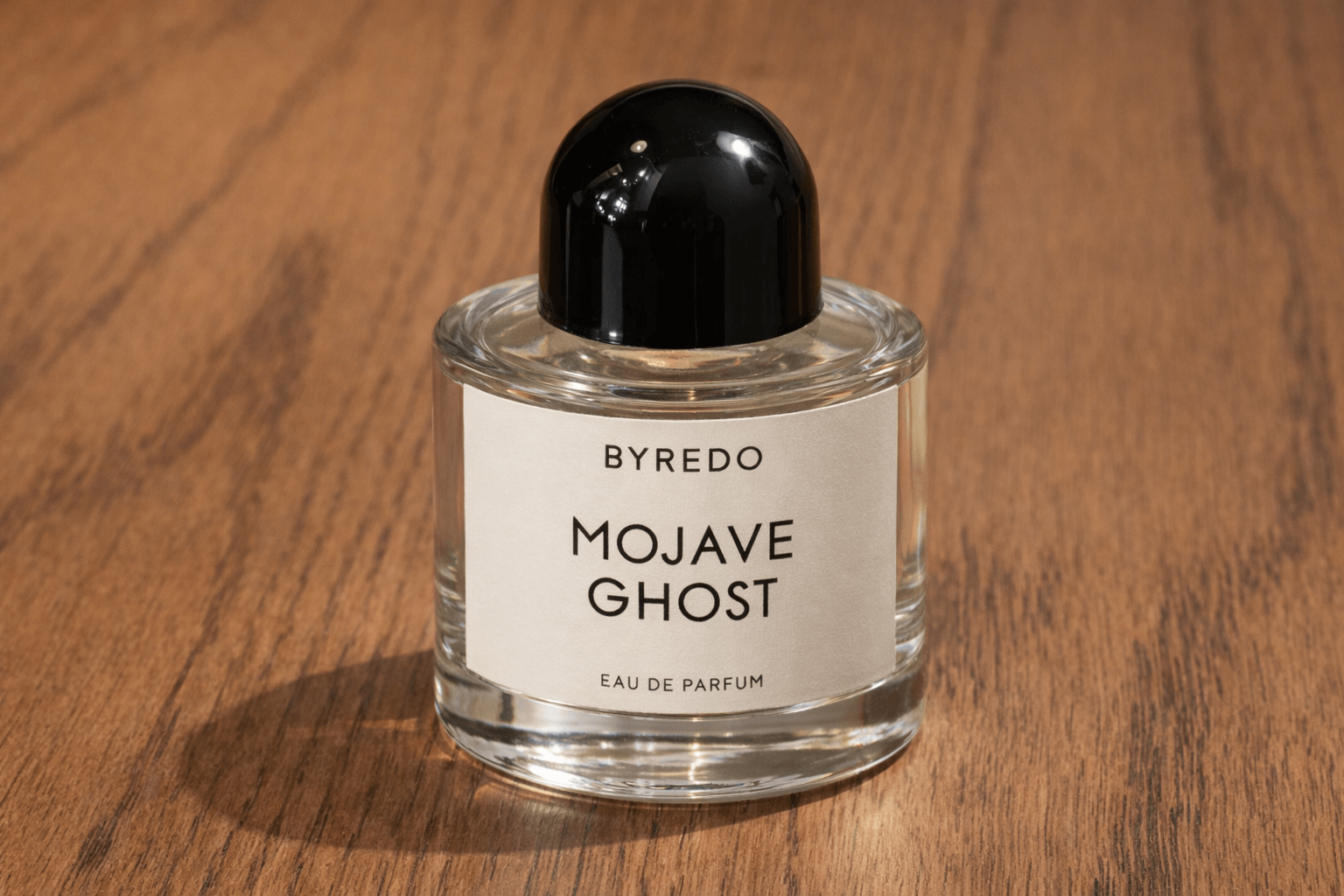 실내 자연광이 비치는 따뜻한 나무 테이블 위에 바이레도 모하비 고스트 향수병이 단독으로 놓여 있다. 향수병은 완전한 원통형의 투명한 유리로 만들어졌으며, 두꺼운 유리 바닥이 아래쪽에서 묵직하게 보인다. 병 위에는 반짝이는 유광 검은색 반구형 뚜껑이 얹혀 있고, 전면에는 크림빛이 도는 오프화이트 종이 질감의 라벨이 붙어 있다. 라벨 중앙에는 &lsquo;BYREDO&rsquo;와 &lsquo;MOJAVE GHOST&rsquo;라는 검은 글자가 정돈된 산세리프 글꼴로 인쇄되어 있으며, 하단에는 작은 크기로 &lsquo;EAU DE PARFUM&rsquo; 문구가 보인다. 전체적으로 광고 촬영이 아닌 일상적인 제품 사진처럼 자연스럽고 차분한 분위기를 전한다.