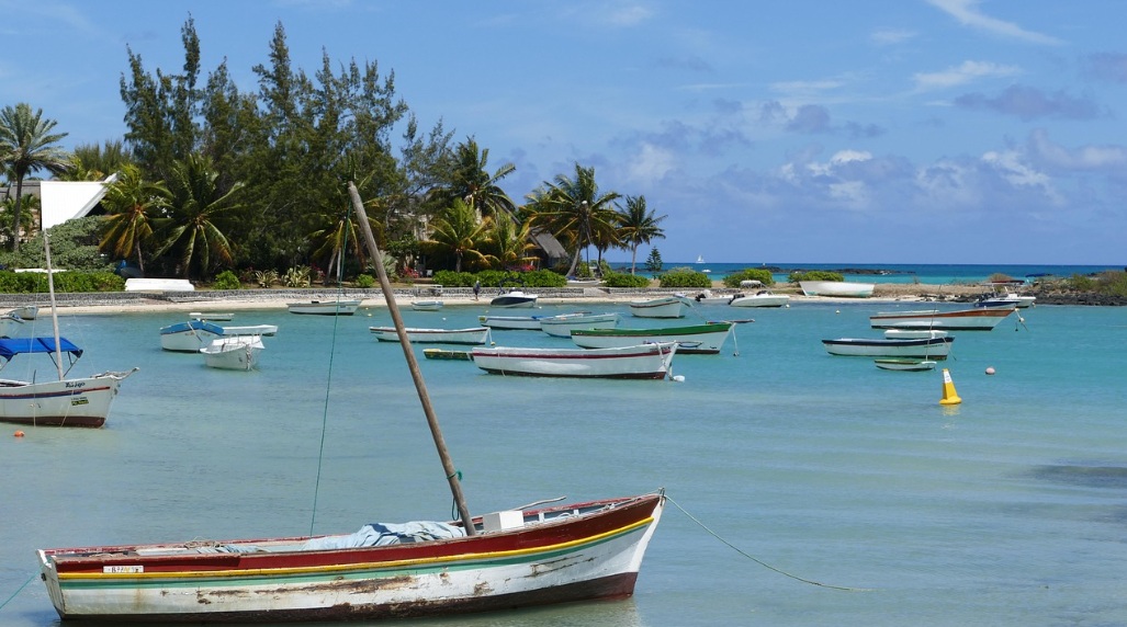 맑고 투명한 에메랄드빛 바다 위에 여러 척의 작은 목선들이 떠 있는 모리셔스의 해변 풍경. 해안선을 따라 야자수와 열대 식물들이 우거져 있으며, 평온하고 이국적인 휴양지의 분위기를 자아냄