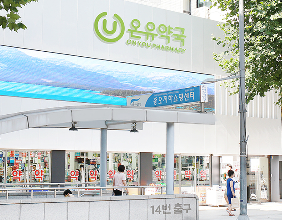 지하철-14번-출구-앞-대형-LED-간판과-흰색-외벽이-돋보이는-온유약국-건물-전경으로-약국-유리창에는-빨간색-약-문구가-반복되어-붙어있고-간판-상단에는-하늘색-종오지하쇼핑센터-안내판과-디지털-파노라마-화면이-설치되어-있으며-나무-그늘과-지나가는-사람들까지-포함된-활기찬-도심-약국-풍경