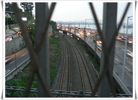 철조망 너머로 보이는 곡선형 철로와 그 옆을 따라 병행하는 강변북로의 풍경. 철로는 중앙에서 멀리 한강을 따라 뻗어 있으며, 오른쪽 도로에는 퇴근 시간대의 차량들로 가득하다. 하늘은 흐릿하게 밝으며, 강변 도시의 바쁜 일상이 느껴지는 장면이다.