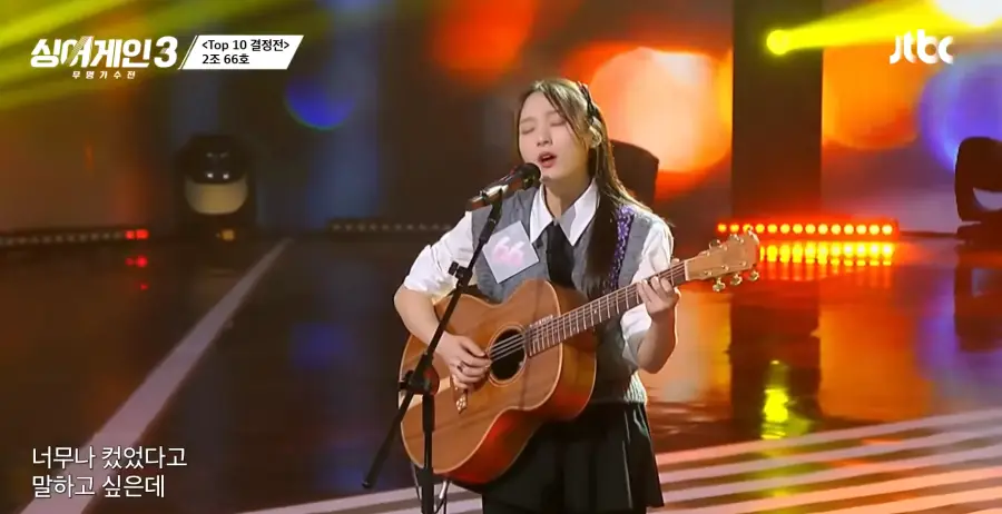 싱어게인3- 검정색 미니스커트에 흰셔츠&amp;#44; 회색 조끼를 입고 검은색 넥타이를 맨체 갈색기타를 들고 긴생머리 양쪽으로 삔을 꽂은채 스탠딩마이크 앞에서 두눈을 감고 노래하는 66호