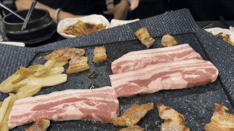 불판 위에 제주산 생오겹살이 올려지고 익기 시작하는 장면. 고기 옆엔 백김치와 구운 고기 조각들이 함께 있다.