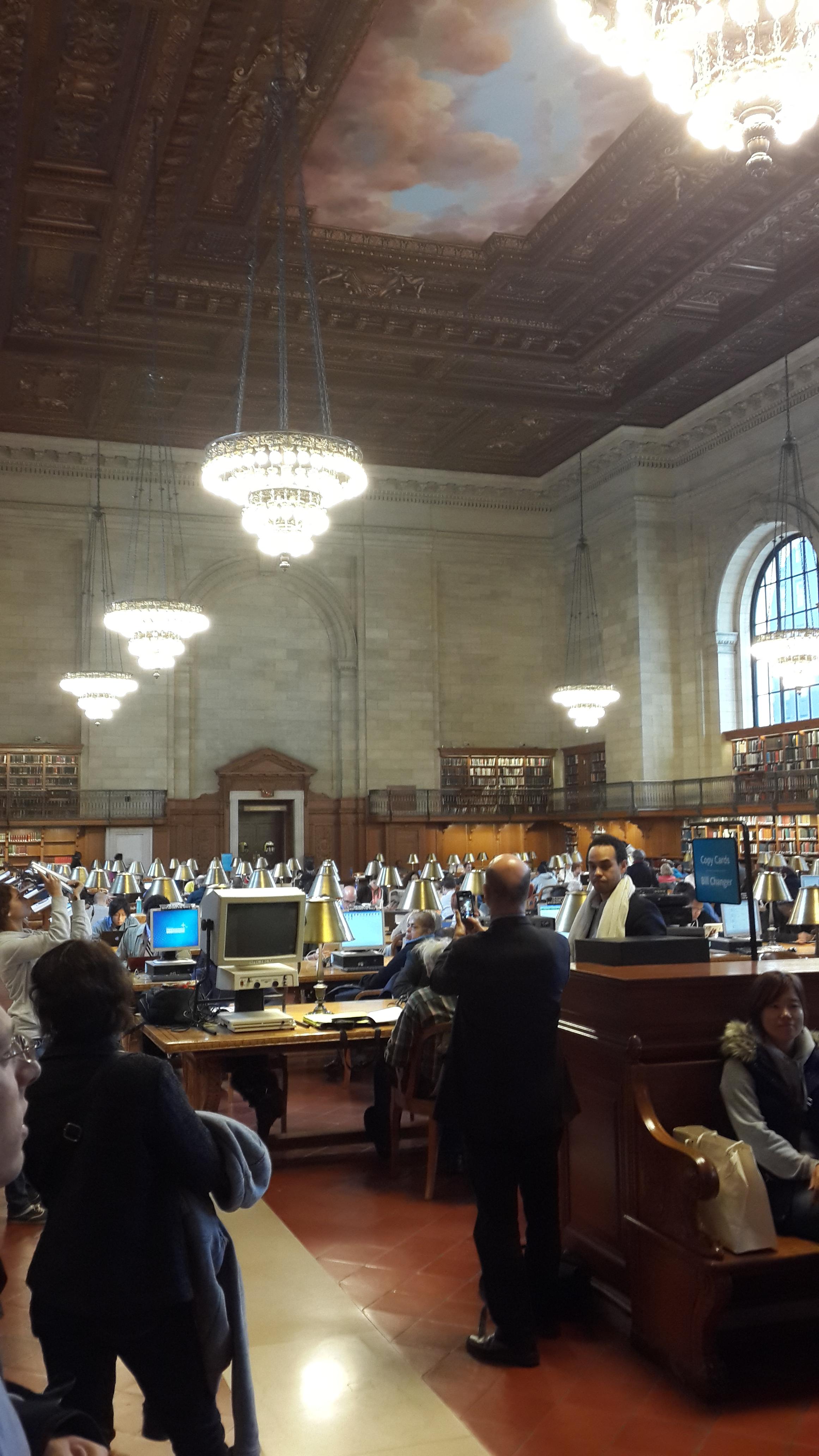 뉴욕 공립도서관 로즈 메인 열람실의 화려한 목재 천장과 구름이 그려진 벽화, 그리고 길게 내려온 금색 샹들리에 조명들.