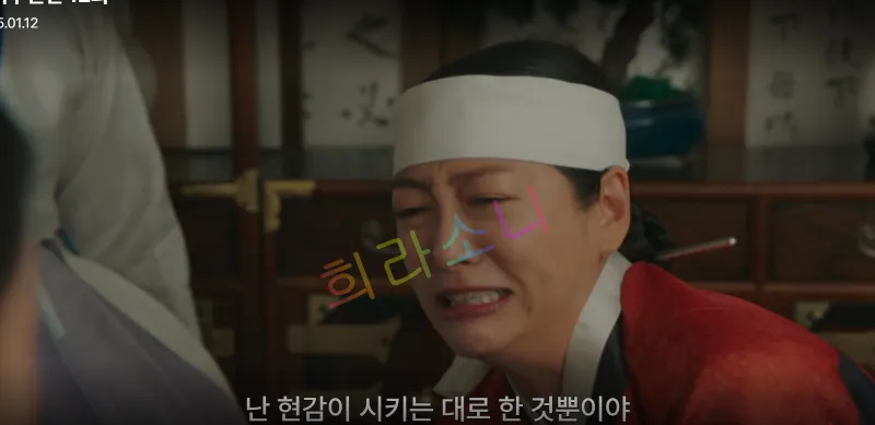 머리에 흰 두건을 두른 홍씨부인이 울먹이며 화난 표정으로 말을 하고 있다. 배경에는 나무 책장이 놓여 있다. 자막 