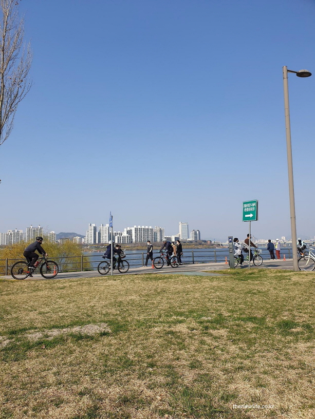 People riding bicycles and walking near the river on a clear day./맑은 날, 강가에서 자전거를 타고 걷는 사람들.