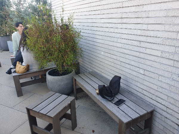 야외 좌석 중 일부 의자에 가방과 핸드폰이 놓여 있다. 화분 뒤 쪽으로 앉아 있는 사람들도 보인다.