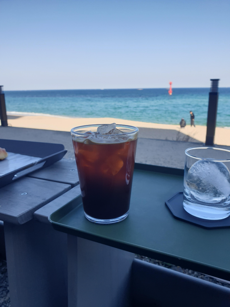 바다가 보이는 테이블 위에 아이스 커피 한 잔과 얼음 컵이 놓여 있다.