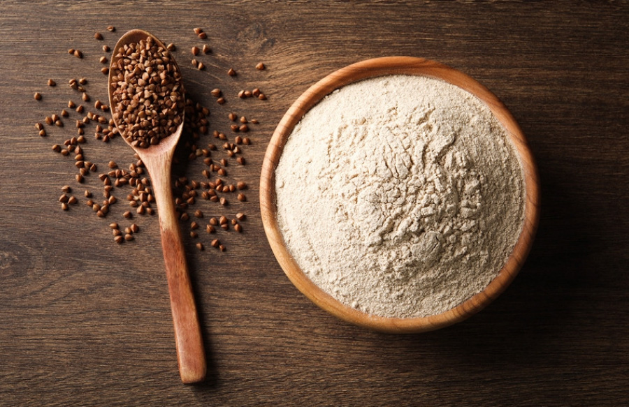 고동색의 바닥 위에 메밀과 메밀을 가득 담은 나무 숟가락을 두고 그 오른쪽에는 메밀가루를 가득 담아둔 고동색의 동그란 나무 그릇을 놓아두고 찍은 사진