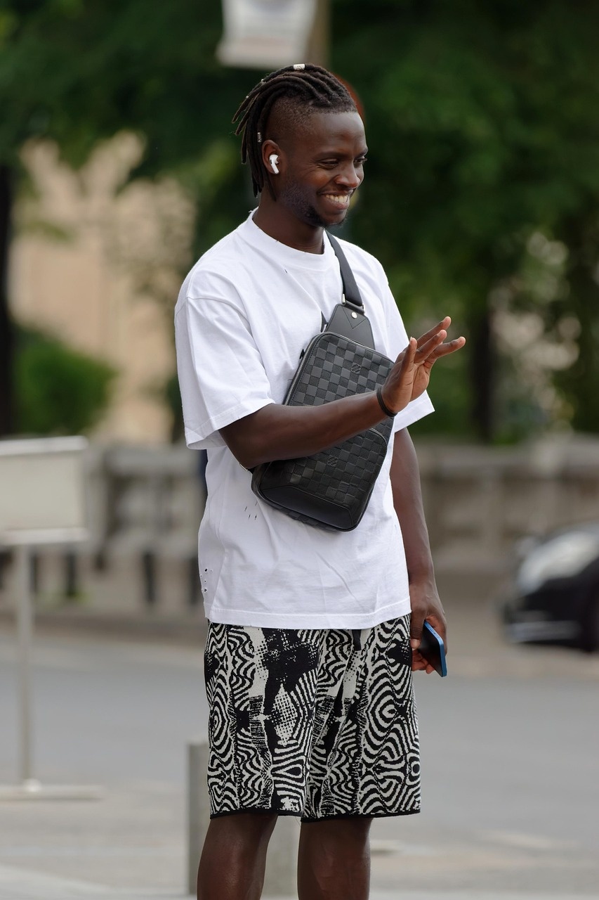 레게머리-남성이-크로스가방을-메고-왼손에는-휴대폰을 들고-오른손으로-인사를-하고있음