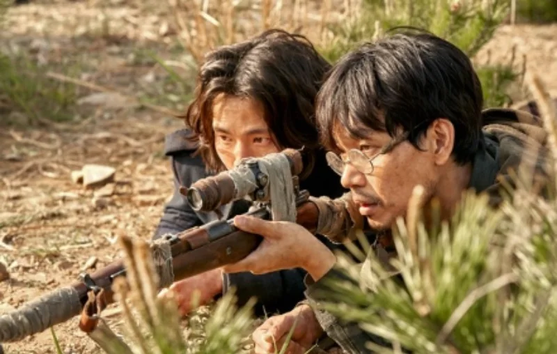 어느 풀숲에 안경을 쓰고 누어 장총으로 누군가를 겨누고 있는넷플릭스 드라마 도적 칼의 소리의 강산군 캐릭터