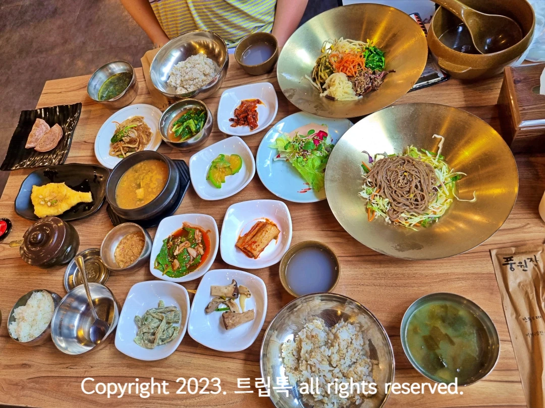 모밀국수&#44; 미역구&#44; 나물&#44; 된장국&#44; 수수부꾸미 등 다양한 반찬과 보리밥이 놋그릇에 담겨있는 식탁