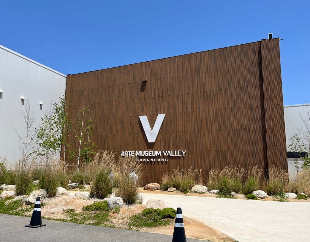 아르떼뮤지엄 강릉 외관 전경, 브라운 우드 패널 벽면에 대형 V 로고와 ARTE MUSEUM VALLEY GANGNEUNG 간판이 보이는 모습