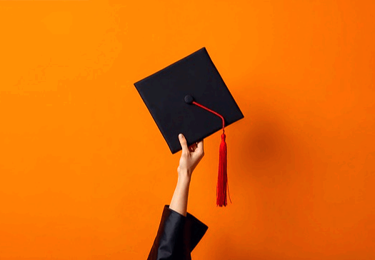 주황색 바탕화면에서 네모난 검정색 학사모를 한 손으로 높이 들고 있다.
