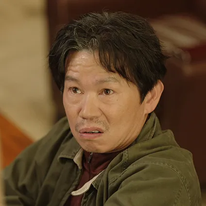 어느 실내를 배경으로 카키색 잠바를 걸치고 누군가를 인상쓰고 있는 반학수