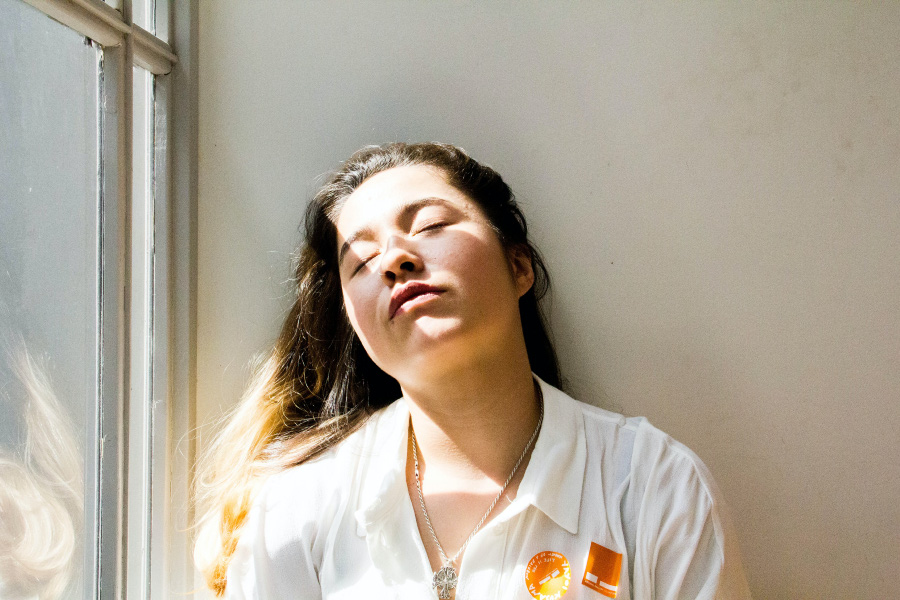 흰색 옷을 입고 있는 머리가 긴 서양 여성이 춘곤증 때문에 나른하고 피곤하여 벽에 기대서 눈을 감고 쉬고 있는 것을 찍은 사진