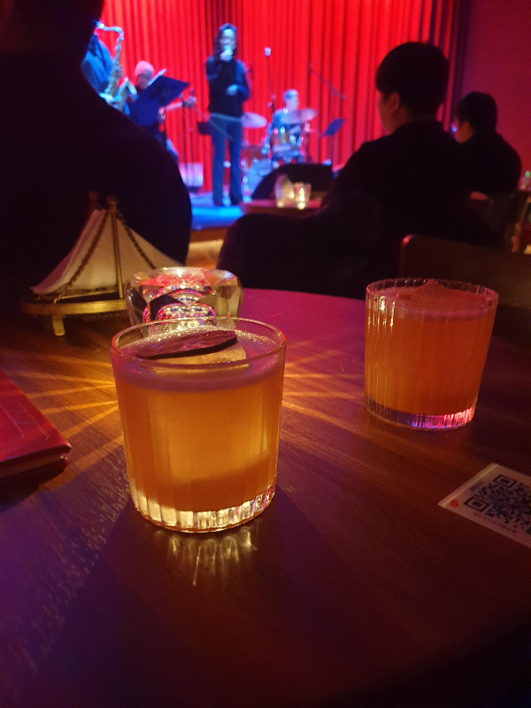 주문한 칵테일 2잔이 테이블에 올려져 있다. 뒤쪽에 공연 중인 하우스 밴드가 보인다.