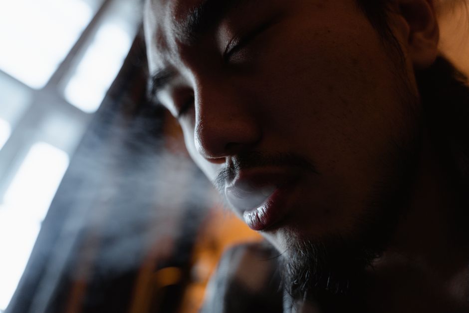 A close-up image of a man exhaling smoke with soft, moody lighting indoors.