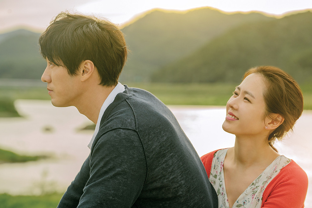 강이 보이는 자연 배경 앞에서 우진과 수아가 등을 맞대고 앉아 있는 장면, 수아가 우진을 바라보며 환하게 웃고 있다