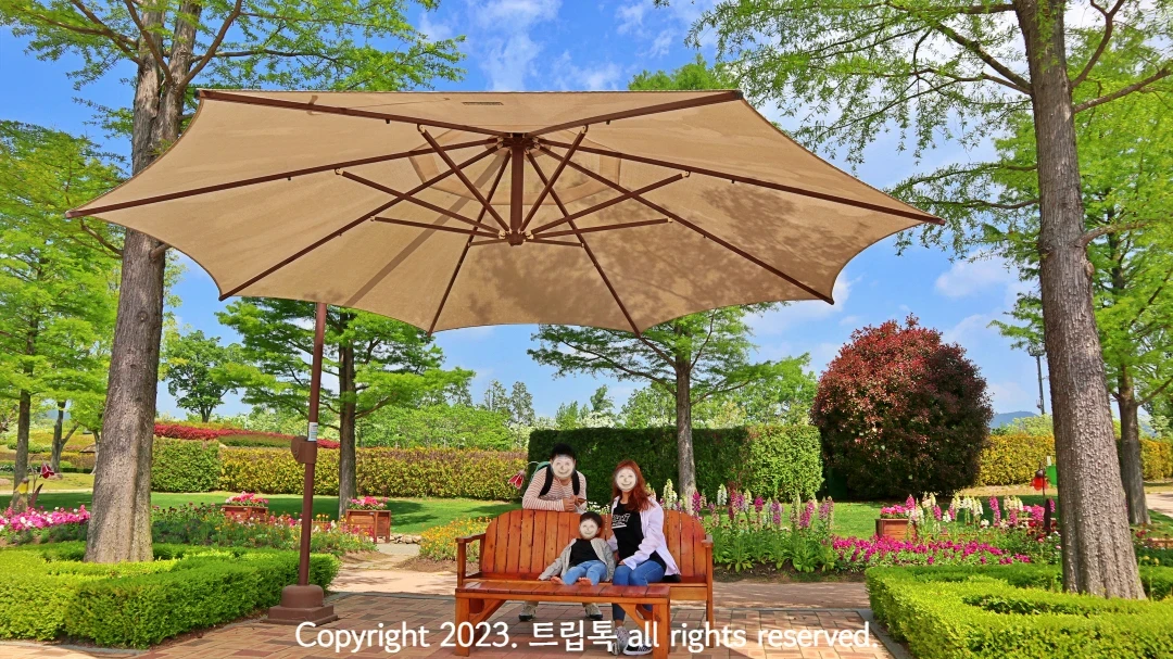 순천만국가정원 내부에 양 옆에 두그루 나무가 있고 가운데 베이지색 파라솔이 펼쳐진 벤치에 세사람이 있다.