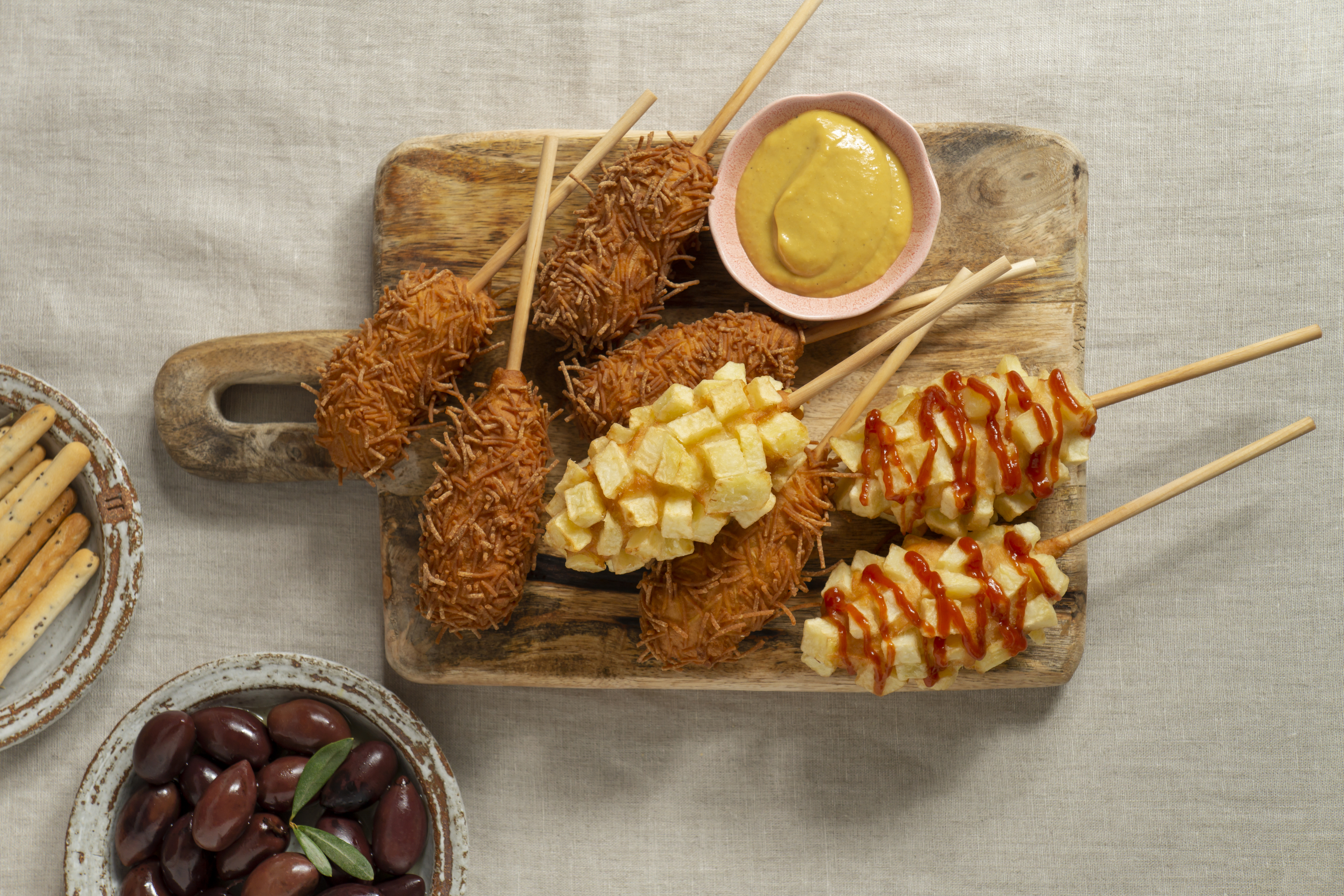 Korean-style corndogs topped with crispy potato cubes and fried noodles, served with mustard dipping sauce on a rustic wooden board.