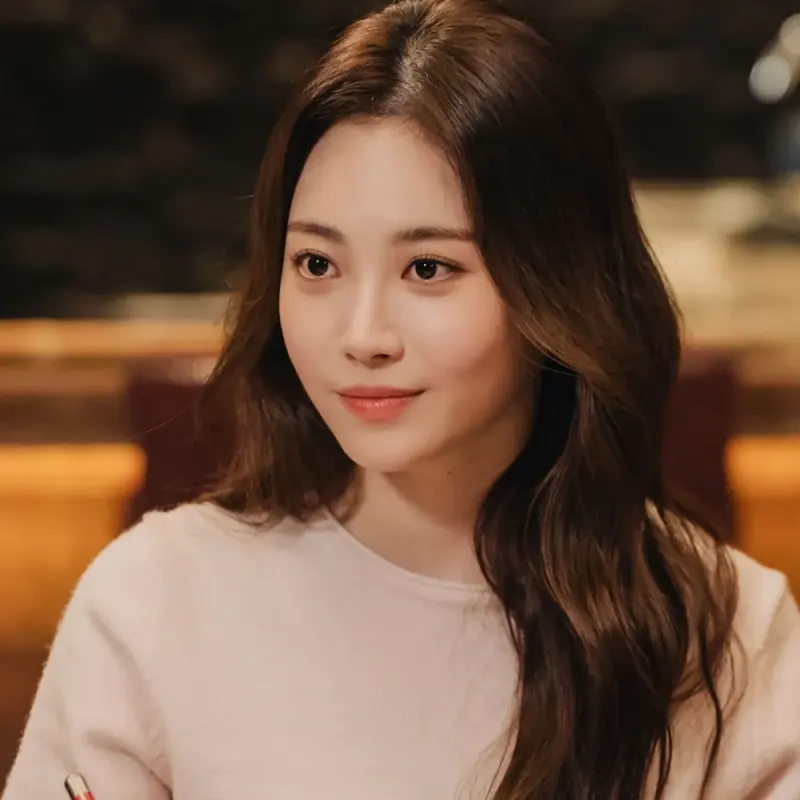 밝은 분홍색 티셔츠에 갈색 긴 머리를 하고 앉아 있는 이 연애는 불가항력의 윤나연