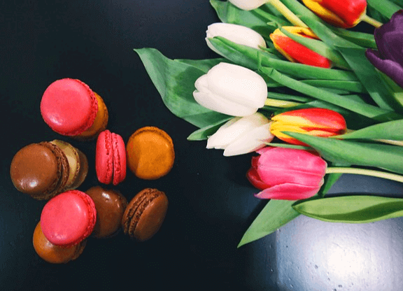 왼쪽엔 연지색 마카롱, 초콜릿마카롱이 여러개 있다. 오른쪽엔 하얀색과 빨간색 튤립꽃들이 있다.