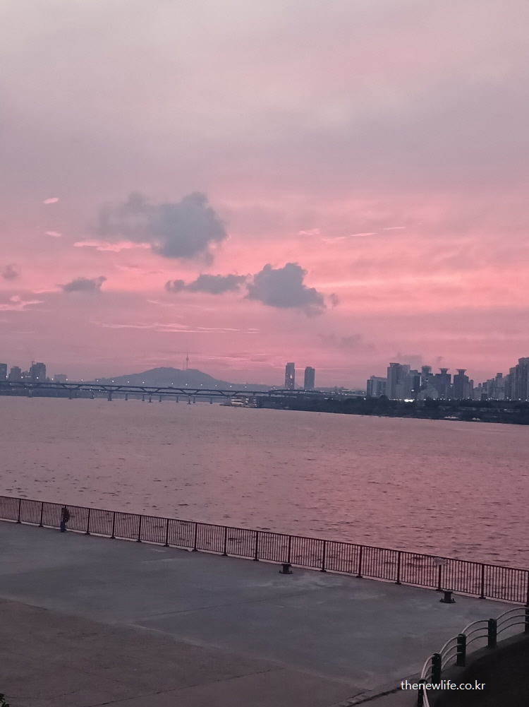 잠실 한강공원 물놀이장에서 한강 너머로 붉게 물든 석양과 남산타워 실루엣이 보이는 감성적인 하늘과 강변