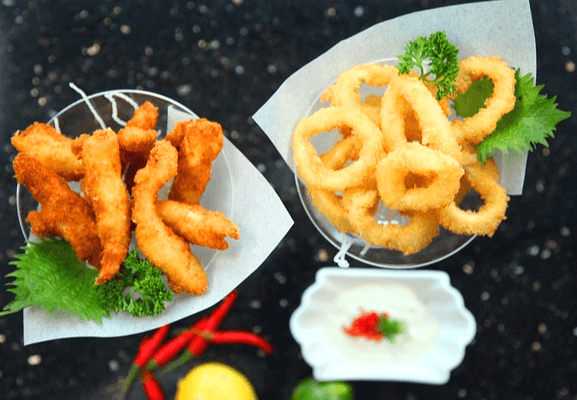 검정 식탁 위에 그릇에 담긴 노릇노릇한 오징어 튀김들이 있다. 긴 몸통 튀김들과 동그란 오징어튀김들이 각각 그릇에 담겨있다.