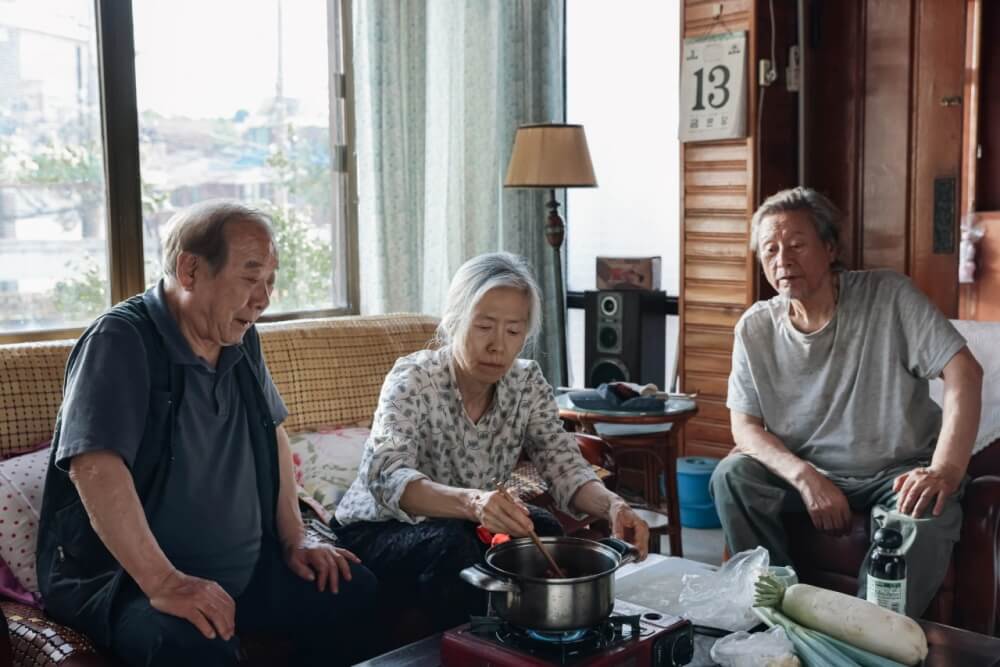 영화 사람과 고기 스틸컷 – 세 노인이 방 안에서 함께 음식을 만드는 잔잔한 일상 장면