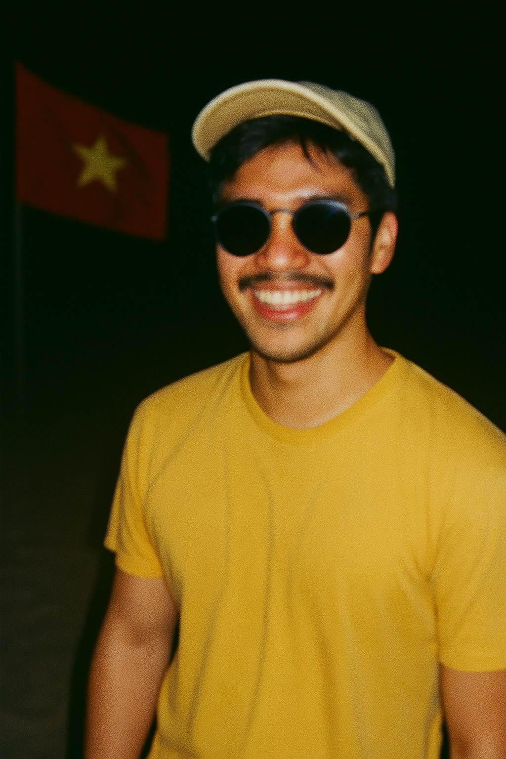 밝은 색 티셔츠에 선글라스, 모자를 쓰고 해변에서 환하게 웃고 있는 남부 베트남 남자, 배경에는 흐릿하게 베트남 국기가 펄럭이고, 밤해변만의 분위기가 느껴진다