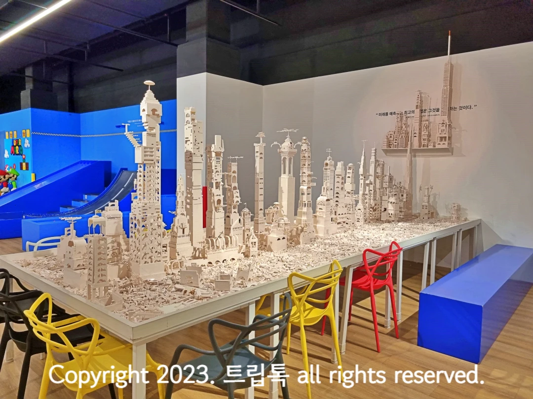 하얀색 브릭으로 20개도 넘는 건물들이 지어져있다. 테이블 옆에는 노랑 검정 빨강 의자가 있다.