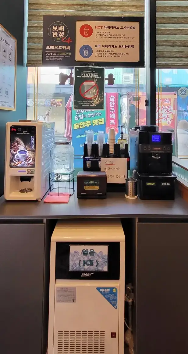 보배반점 - 나무 테이블 위 흰색 믹스커피 자판기 옆 물이 가득 담긴 투명 생수병 옆 종이컵과 플라스틱 컵이 가득 쌓인 컵 홀더 옆 아메리카노 커피 자판기와 테이블 아래 하얀색 큰상자의 얼음만드는 기계