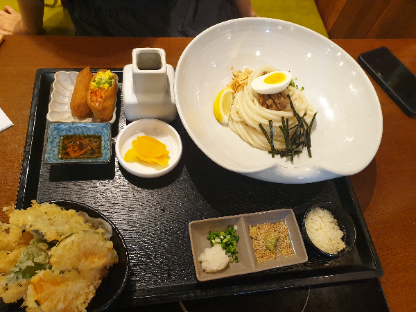 큰 그릇에 냉우동이 있고 2개의 유부초밥과 모듬 튀김 그리고 소스들이 보인다.