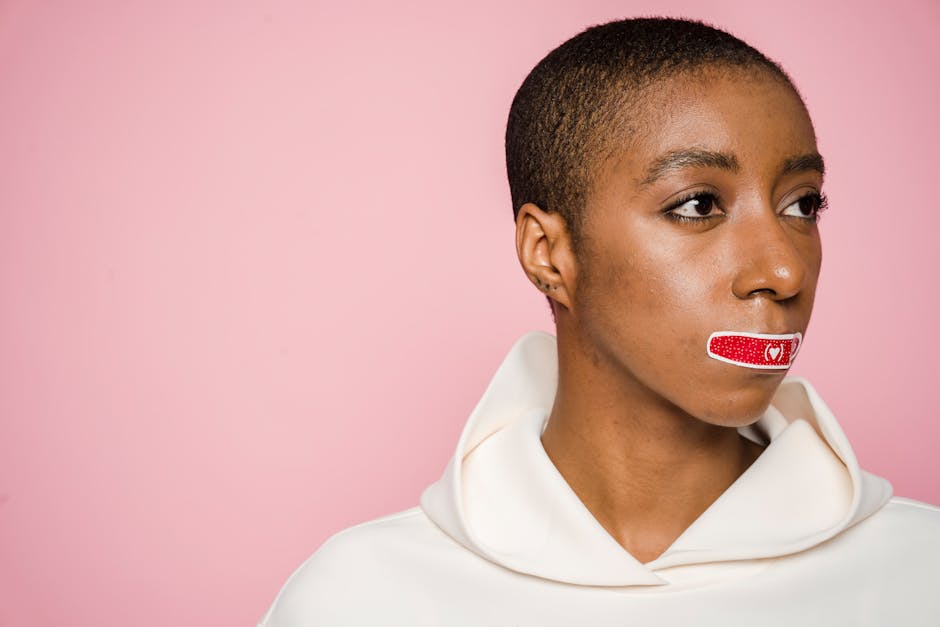 Empowered African American woman with Band-Aid on lips, symbolizing silence and awareness on a pink background.