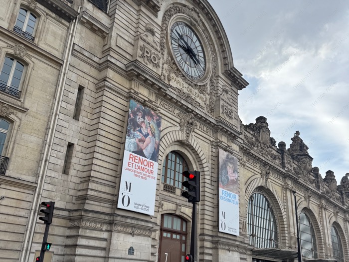 The grand facade of Musee d'Orsay in Paris with a large exhibition banner for Renoir and Love under a cloudy sky⁠