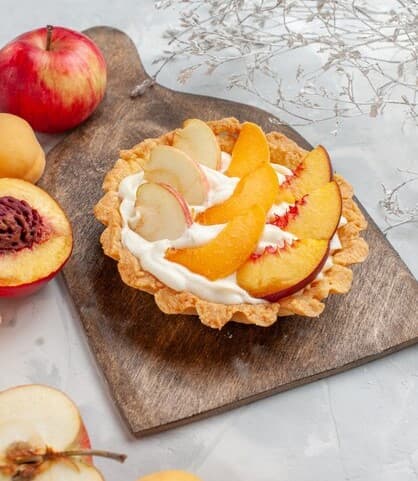 나무 도마 위에 올려진 과일 타르트. 흰 크림 위에 얇게 슬라이스 된 사과, 복숭아, 살구가 가지런히 올려져 있다