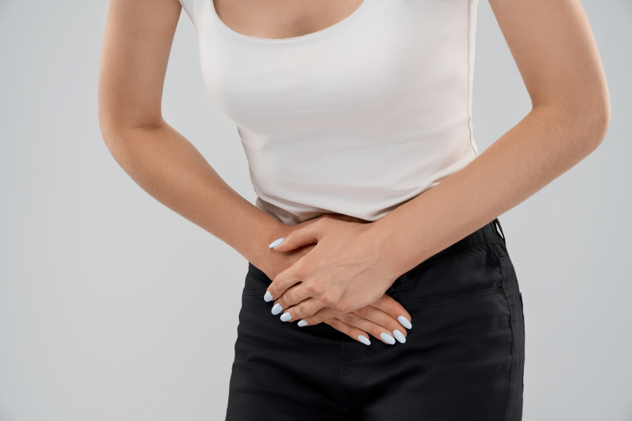 흰색 나시티와 검은 치마를 입은 젊은 여성이 생리통을 참기 위해 양쪽 손을 아랫배에 대고 통증을 참고 있는 것을 찍은 사진