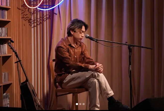 싱어게인3- 갈색 커튼을 배경으로 베이지색 바지에 갈색 셔츠를 입고 의자에 앉아 스탠딩 마이크를 앞에두고 땅을 보며 앉아있는 47호