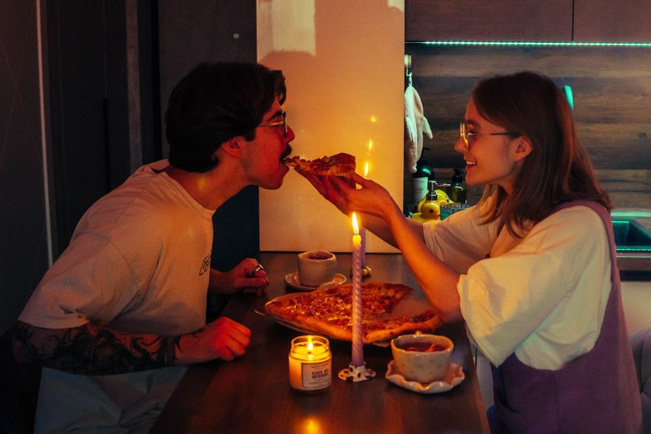 Couple shares a romantic candlelit pizza dinner in a cozy kitchen setting.