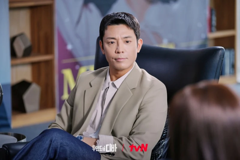 어느 사무실을 배경으로 베이지색 자켓을 입고 앉아 있는 무인도의 디바 이서준 캐릭터