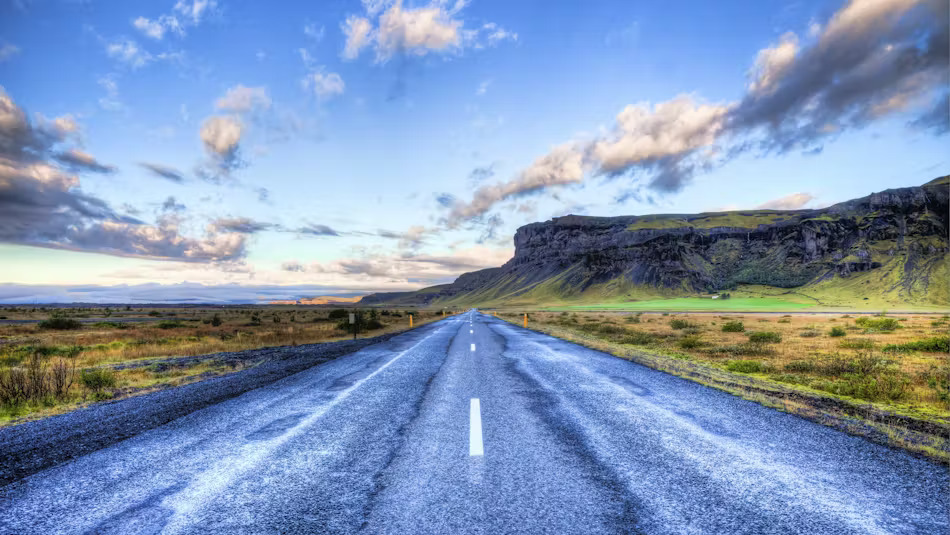 아이슬란드의 링로드(1번 국도)를 보여주고 있습니다. 포장된 도로가 드넓은 아이슬란드 평원을 가로지르며 지평선까지 쭉 뻗어 있고, 오른쪽으로는 웅장한 절벽 지형이 보입니다. 푸른 하늘에 흰 구름이 떠있는 전형적인 아이슬란드의 여름 풍경입니다.