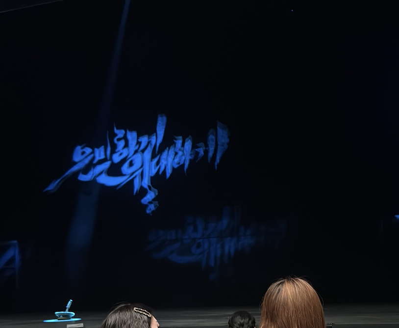 뮤지컬 공연장 객석에서 바라본 무대 스크린 이미지. 짙은 파란색의 역동적인 한글 캘리그라피가 무대 어둠 속에 빛나고 있으며, 그 아래로 흐릿한 잔상(vibration)이 남아 공연의 전율과 여운을 시각적으로 표현하고 있다