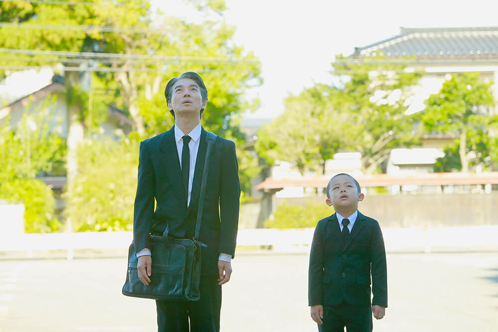 미조구치가 아들과 함께 양복을 입고 묘비석 방문판매를 다니다 위를 쳐다보고 있는 모습