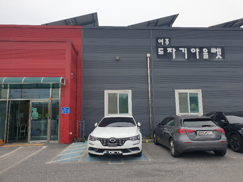 회색 조립식 판넬 건물에 붉은색 입구가 포인트가 특징인 건물앞에 차량들이 주차 되어 있다.