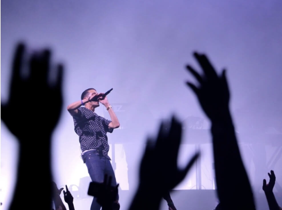 남성이 무대 위에서 노래하고 있다. 관중들은 손을 올리며 환호하고 있다