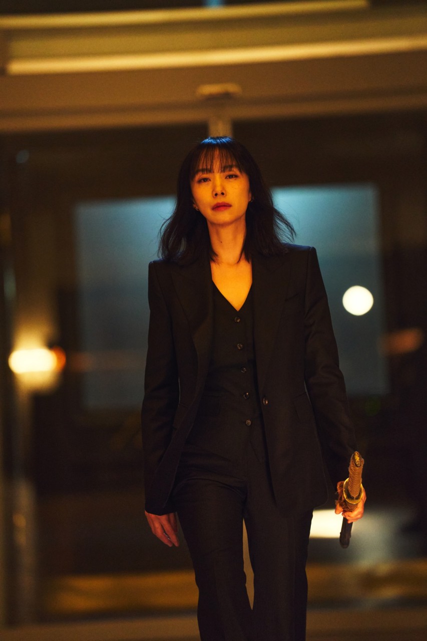주인공 복순이 검은 수트에 어두운 표정을 하고 왼손에 칼을 쥐고 어디론가 가고 있다.
