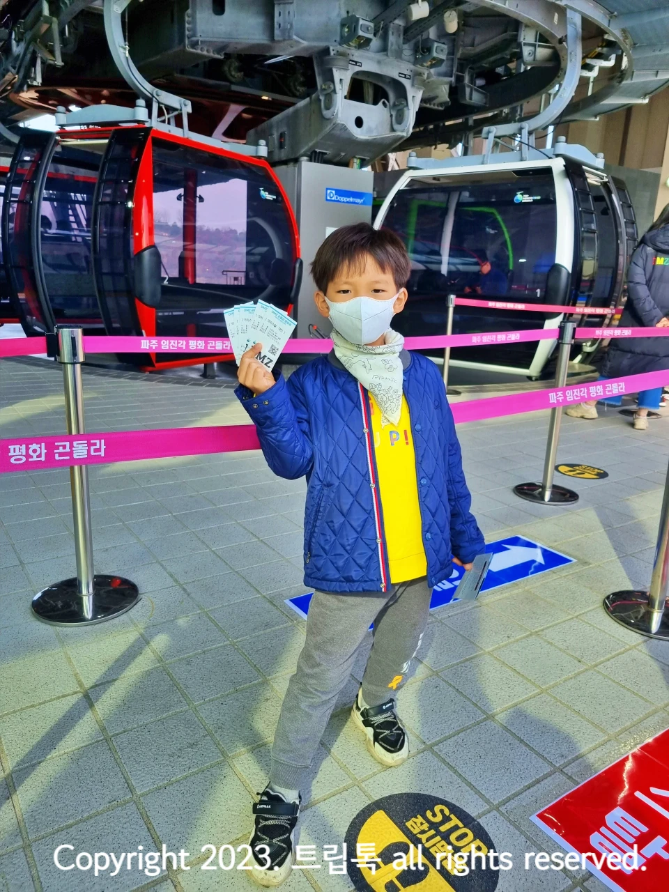 빨간색 하얀색 파주 임진각 곤돌사 사이에 파란 잠바를 입은 아이가 티켓을 들고 있다.
